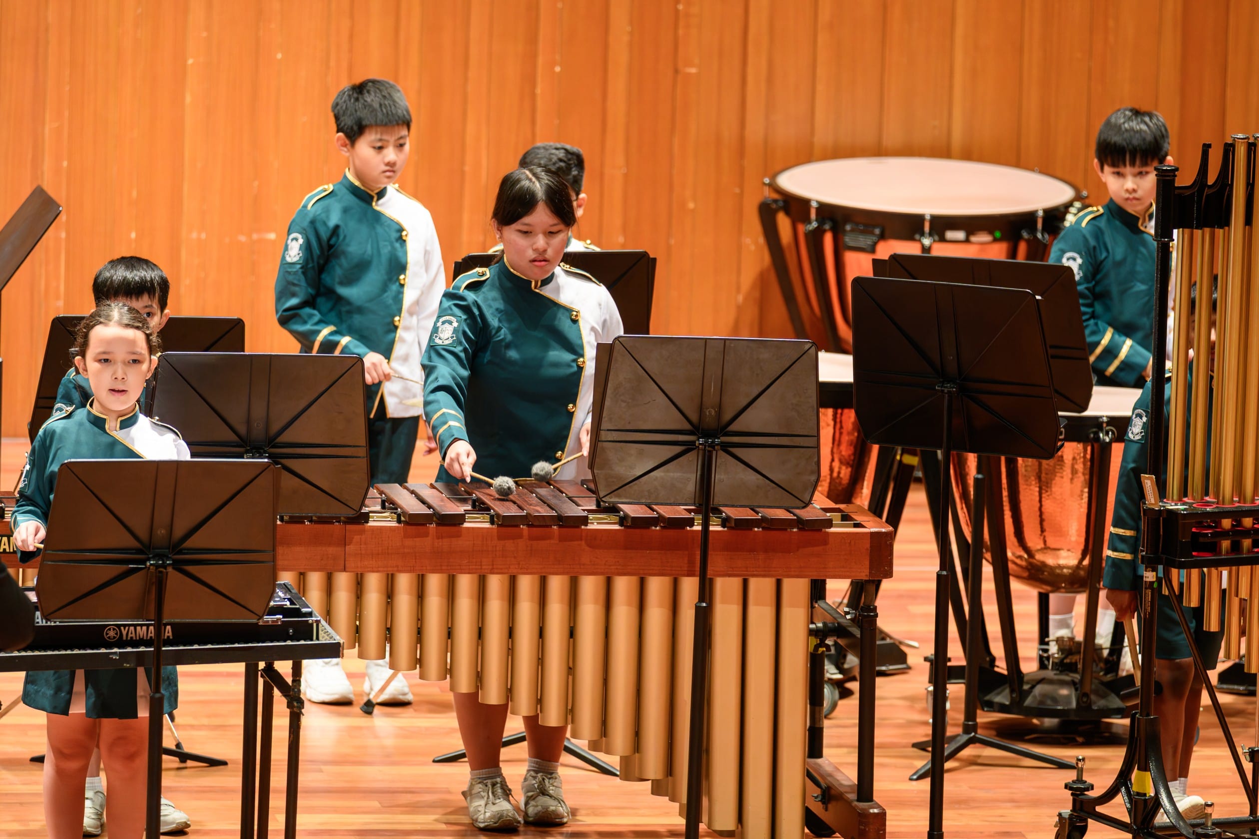School band students in uniform performing a percussion ensemble on a concert stage, with a student playing the marimba as the focal point, accompanied by bandmates at a keyboard and other percussion instruments in the background.